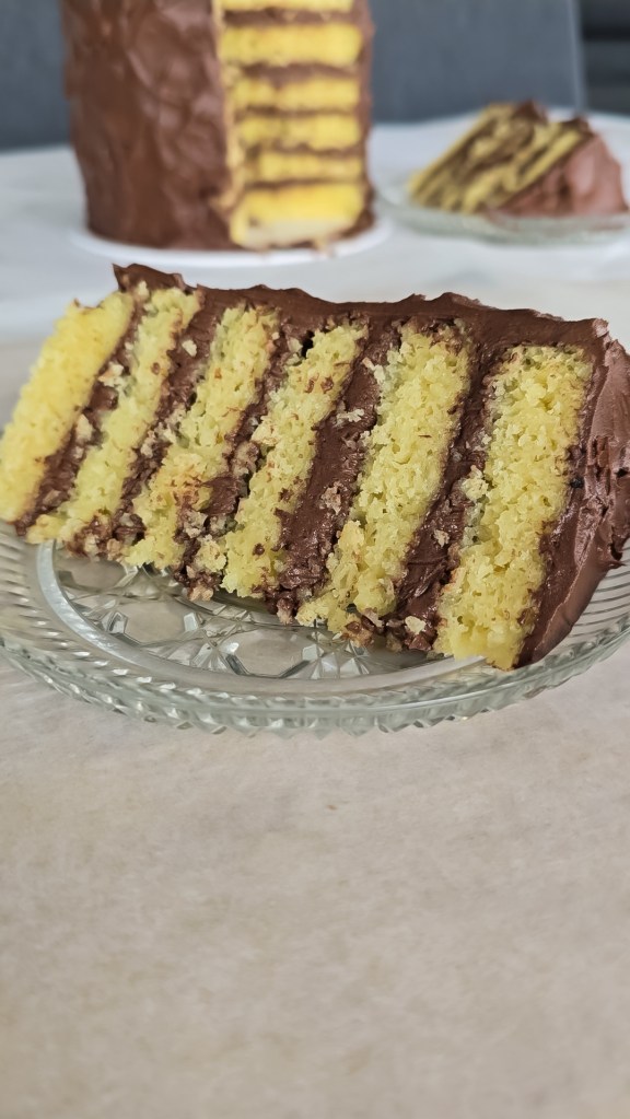 A slice of yellow cake with chocolate buttercream on a plate with the rest of the cake in the background.